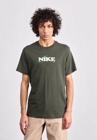 Oliven grøn bomulds t-shirt med korte ærmer, med "NIKE" logo i hvid og lys grøn tekst. Klassisk rund halsudskæring og afslappet pasform.