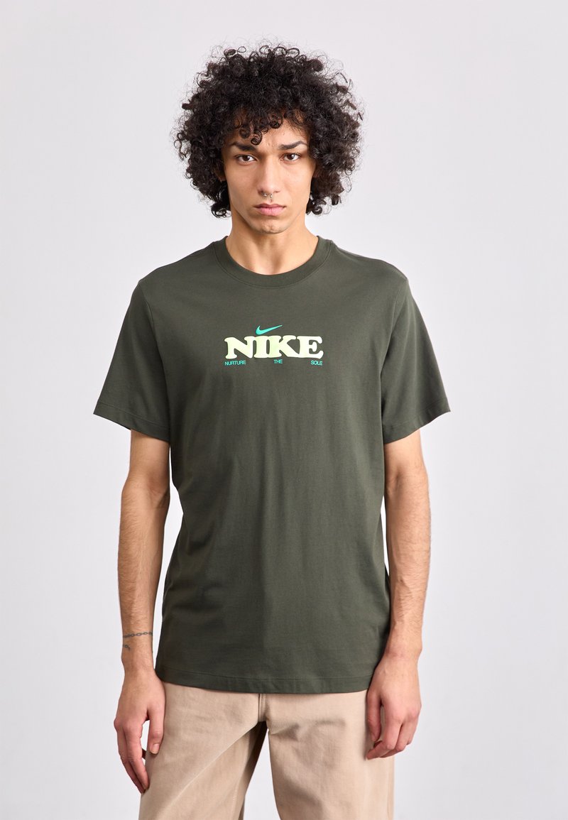 Oliven grøn bomulds t-shirt med korte ærmer, med "NIKE" logo i hvid og lys grøn tekst. Klassisk rund halsudskæring og afslappet pasform.