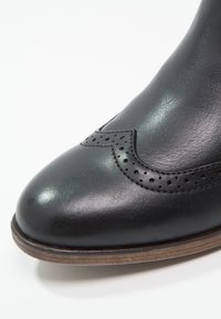 Stivale alla caviglia in pelle nera con punta arrotondata, caratterizzato da perforazioni decorative e un design scalloped lungo il lato, e una suola marrone.