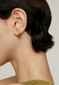 Pendiente de aro dorado con un diseño floral, usado en la oreja izquierda. Cabello corto y oscuro y piel texturizada visibles en un primer plano. Ropa de color verde oliva.