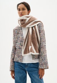Eșarfă cu model maro și alb așezată peste o jachetă de tweed cu pete colorate, combinată cu un top alb și blugi blue.