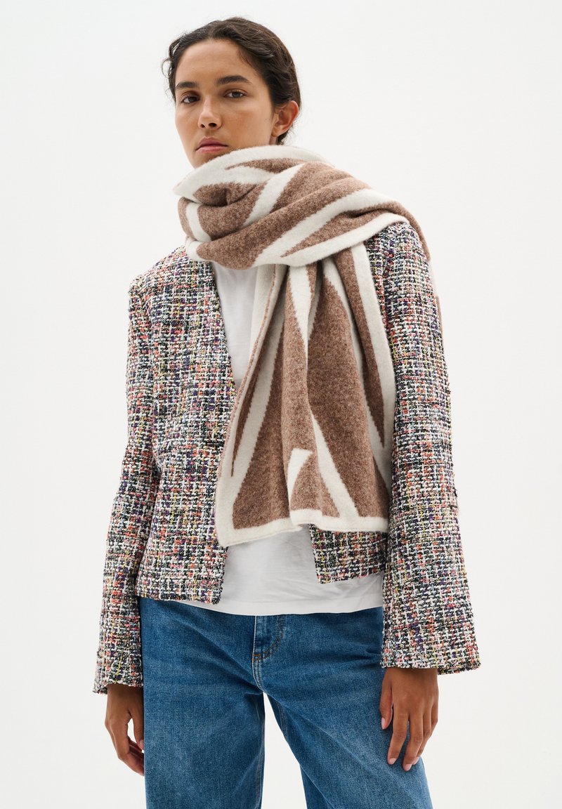 Eșarfă cu model maro și alb așezată peste o jachetă de tweed cu pete colorate, combinată cu un top alb și blugi blue.