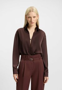 Blusa lunga in satin marrone con scollatura a V profonda e colletto abbottonato, abbinata a pantaloni a vita alta con cintura e pieghe.