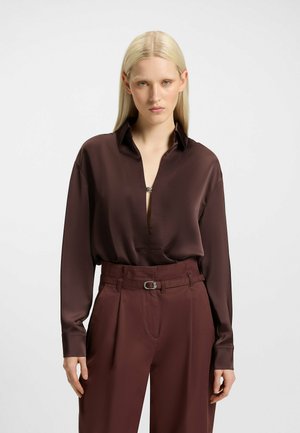Blusa lunga in satin marrone con scollatura a V profonda e colletto abbottonato, abbinata a pantaloni a vita alta con cintura e pieghe.