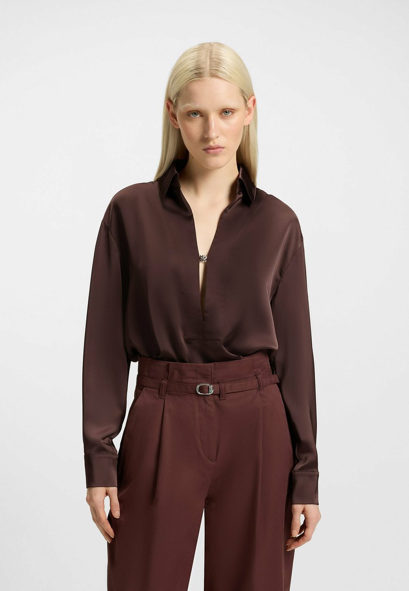 Blusa lunga in satin marrone con scollatura a V profonda e colletto abbottonato, abbinata a pantaloni a vita alta con cintura e pieghe.