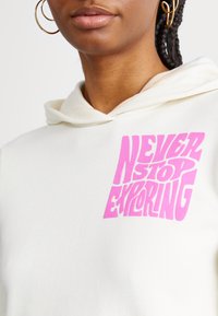 Gros plan d'une personne portant un sweat à capuche blanc avec un texte rose indiquant « NEVER STOP EXPLORING » et des boucles d'oreilles créoles dorées.