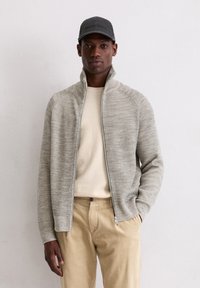 Lichtgrijze ritsjack met ribbeltextuur, gedragen over een crèmekleurige trui en gecombineerd met beige corduroy broek. Grijze baseballpet als accent.