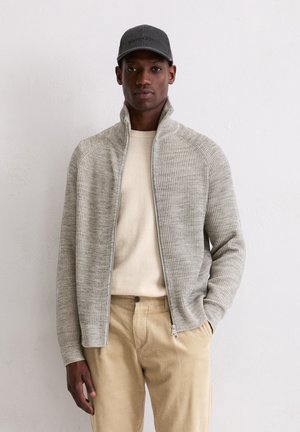 Lichtgrijze ritsjack met ribbeltextuur, gedragen over een crèmekleurige trui en gecombineerd met beige corduroy broek. Grijze baseballpet als accent.