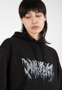 Black hoodie made of soft fabric featuring a drippy silver graphic design on the front. It has a drawstring hood and relaxed fit.