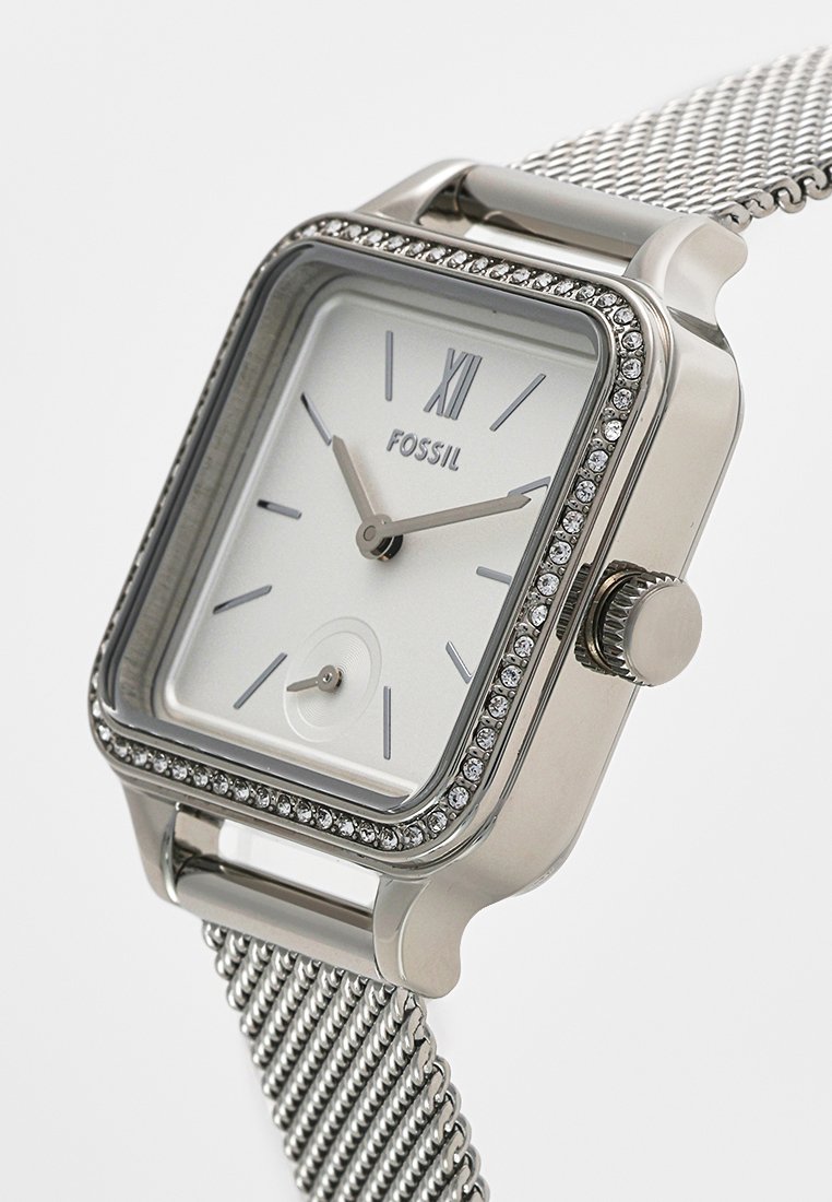 Srebrny kwadratowy zegarek z białą tarczą, rzymskimi cyframi i kryształowymi akcentami na bezelu. Posiada siateczkowy pasek ze stali nierdzewnej i koronkę.