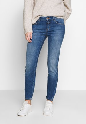 Jeans de denim azul con un ajuste slim, cintura de altura media y un ligero desgaste. Combinados con zapatillas blancas y un suéter beige.