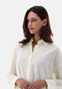 Blusa leggera gialla chiara, trasparente, con texture a pois bianchi, chiusura frontale con bottoni e collo; maniche lunghe con una vestibilità rilassata.