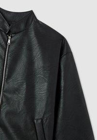 Giacca di pelle nera con collare alto e zip argentata, caratterizzata da una texture liscia e una vestibilità ampia, con dettagli di design minimalisti.