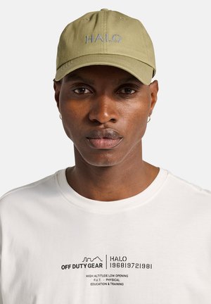 Mann mit mittel-dunkler Haut, der eine hellolivgrüne Mütze mit der Aufschrift "HALO" und ein weißes Hemd mit schwarzer Aufschrift und Grafik "OFF DUTY GEAR" trägt.