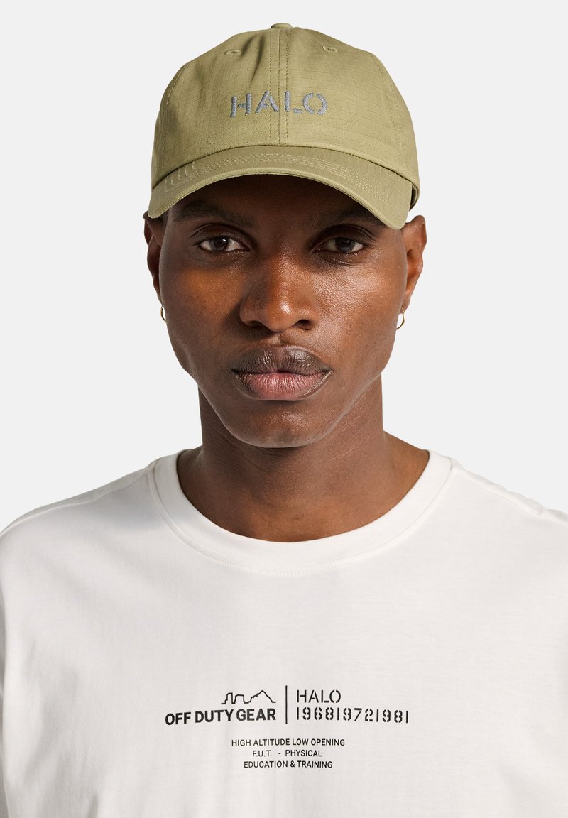 Mężczyzna o średnio-ciemnej karnacji, noszący jasnooliwkową czapkę z napisem "HALO" oraz białą koszulę z czarnym napisem i grafiką "OFF DUTY GEAR".