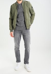 Olivgrüne Bomberjacke über einem grauen Strickpullover, kombiniert mit grauen Jeans und weißen Sneakers. Die Jacke hat eine Zip-Tasche am Ärmel.