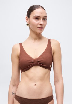 Nő barna, mintás bikiniben, amelynek közepén csomó részlet található, nagy, csillag alakú arany fülbevalókkal és egy kis kereszt tetoválással a karján.