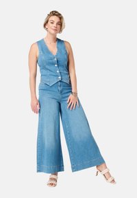 Denim vest met een V-hals en vijf knopen, gecombineerd met wijde denim broek. Lichtblauwe kleur en een gladde textuur. Witte gespenhakken.