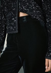 Mustast litritega jakk tekstureeritud pinnaga, mida täiendavad mustad sametpüksid, millel on lamedad nupud ja küljetaskud.