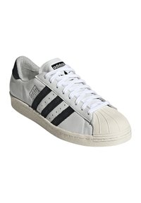 Sneaker in pelle bianca con dettagli neri, tre strisce nere, punta in gomma e lacci bianchi. Design testurizzato e aderente con dettaglio del logo.