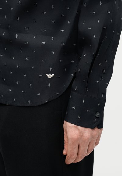 Μαύρο πουκάμισο με μακριά μανίκια, με μικρό μοτίβο από λευκά λογότυπα, μανσέτες με κουμπιά και ένα μεγαλύτερο λευκό λογότυπο κοντά στη μέση, φορεμένο με μαύρο παντελόνι.