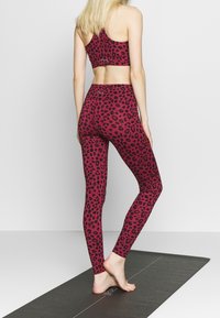 Sportovní podprsenka a legíny v růžovém leopardím vzoru, se střihovým designem a hladkou texturou. Nosené na boso na jogovém koberci.