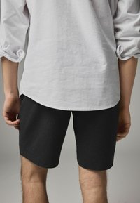 Pantaloni scurți negri cu o textură netedă, având un croi ajustat. Combinați cu o cămașă albă, cu un croi lejer, manșetele fiind desfăcute.