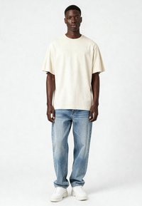 Gaiši bežs īsām piedurknēm T-krekls ar brīvu piegriezumu, kombinēts ar gaiši ziliem taisnajiem džinsiem un baltajiem masīvajiem kedām. Gluda auduma tekstūra.