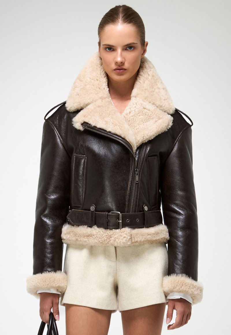Bruine leren jas met een fluffy beige sheepskin kraag en manchetten, met een ritssluiting aan de voorkant, twee zakken en een ontwerp met een ceintuur in de taille.