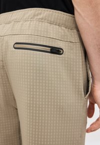 Κοντινό πλάνο μπεζ καρό παντελονιού με οριζόντια τσέπη στο πίσω μέρος με φερμουάρ και ορατή πλαϊνή τσέπη, φορεμένο από άτομο με μαύρο πουκάμισο.