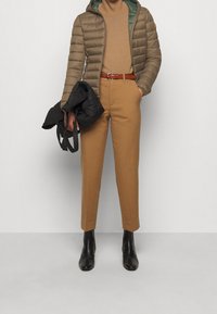 Henkilö, joka on pukeutunut beigen väriseen kaula-aukolliseen puseroon, vaaleanruskeisiin housuihin, jossa on ruskea vyö, vihreäpintaiseen ruskeaan untuvatakkiin, mustiin nilkkabootseihin ja kantaa mustaa käsilaukkua.