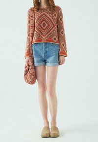 Frau, die einen orangefarbenen gemusterten Pullover trägt, passend zu einer Umhängetasche, Denim-Shorts und beigen Slippern, steht vor einem schlichten Hintergrund.