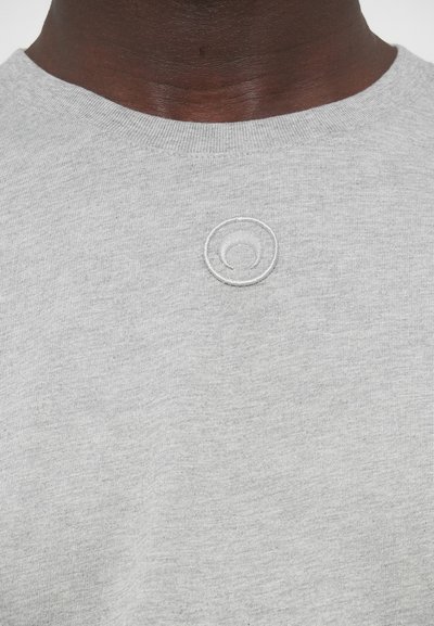 Sivé bavlnené tričko s okrúhlym výstrihom, na hrudi má malú bielu kruhovú výšivku. Tkanina má hladkú, rovnomernú textúru.