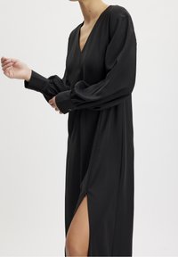 Vestido negro de manga larga en tejido suave, con escote en V, mangas fruncidas y una alta abertura lateral con detalle de cremallera.