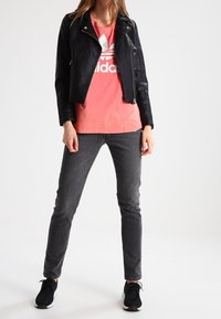 Kvinna i svart läderjacka, korallfärgad Adidas-t-shirt, mörka skinny jeans och svarta sneakers som står mot en neutral bakgrund.