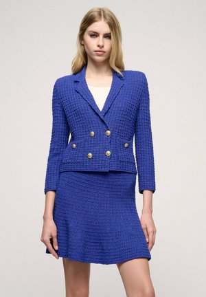 Blue woven blazer with a structured fit, double-breasted design, and gold buttons. Paired with a matching flared skirt in similar material.