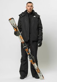 Schwarze Skijacke und Hose mit strukturierter Oberfläche, die Skier mit orangefarbenen Akzenten und Metallbindungen hält. Handschuhe vervollständigen das Outfit.
