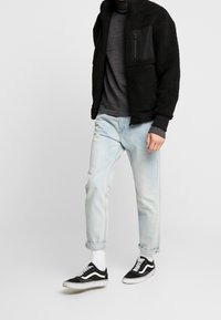 Mustat fleece-takit tekstuuripinnalla ja vetoketjullinen tasku, harmaa neule, vaaleansiniset farkut ja mustat Vans-kengät valkoisilla yksityiskohdilla.