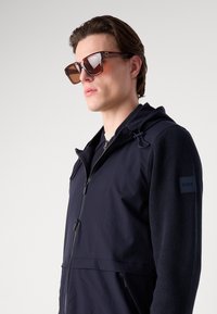 Navy hooded jacket with zip closure, softshell body, and knit sleeves. Brown square sunglasses. Black logo patch on sleeve.