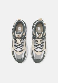 Baskets de sport comprenant une combinaison de matériaux verts, beiges et gris, avec une semelle blanche texturée et des lacets noirs.