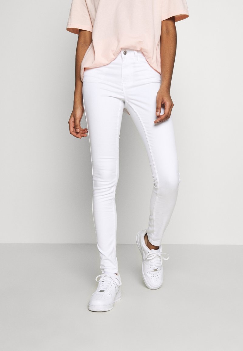 Personne portant un jean skinny blanc, des baskets blanches et un t-shirt rose clair à manches courtes, debout devant un fond uni et clair.