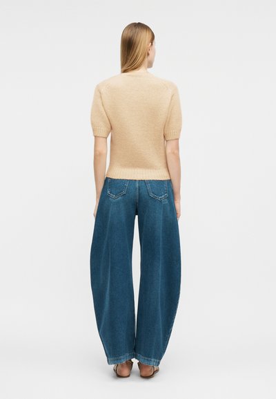 Γυναίκα με ίσια ξανθά μαλλιά, φοράει μπεζ πουλόβερ με κοντά μανίκια και μπλε τζιν παντελόνι με φαρδιά μπατζάκια, στέκεται ξυπόλυτη και γυρίζει την πλάτη της.
