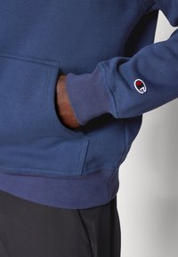 Felpa blu navy con polsini e orlo a costine, dotata di una tasca frontale e un piccolo logo ricamato sulla manica. Texture morbida, design casual.