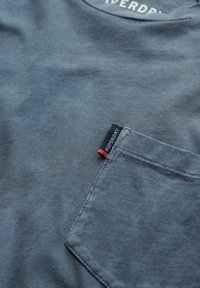 Camiseta de algodón gris con cuello redondo, que presenta un bolsillo frontal y una etiqueta de logo visible en azul y naranja en la costura del bolsillo.