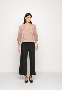 Blusa em rosa pálido com um decote V profundo e mangas transparentes texturizadas, adornada com bordados florais. Combinada com calças pretas de cintura alta.