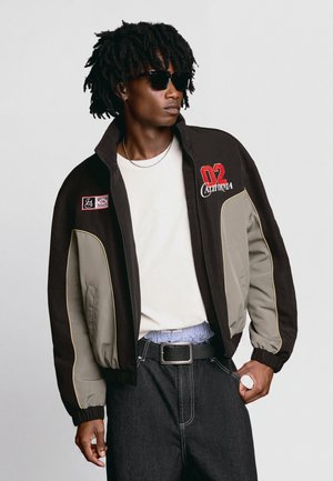 Junger Mann mit Sonnenbrille, der eine schwarz-graue Jacke mit dem Schriftzug "02 California", ein weißes Hemd, schwarze Jeans und eine silberne Kettenhalskette trägt.