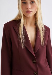 Giovane donna con lunghi capelli biondi che indossa un blazer su misura di un profondo bordeaux con due bottoni, senza camicia visibile sotto.