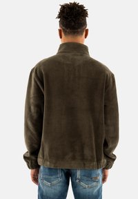 Suéter de pana verde olivo con cuello alto y textura acanalada, con puños elásticos y un corte relajado, combinado con jeans azules.