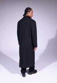 Long manteau noir avec un design ajusté, en matériau de laine, et une fente au dos. Associé à un pantalon noir et des bottes noires imposantes.