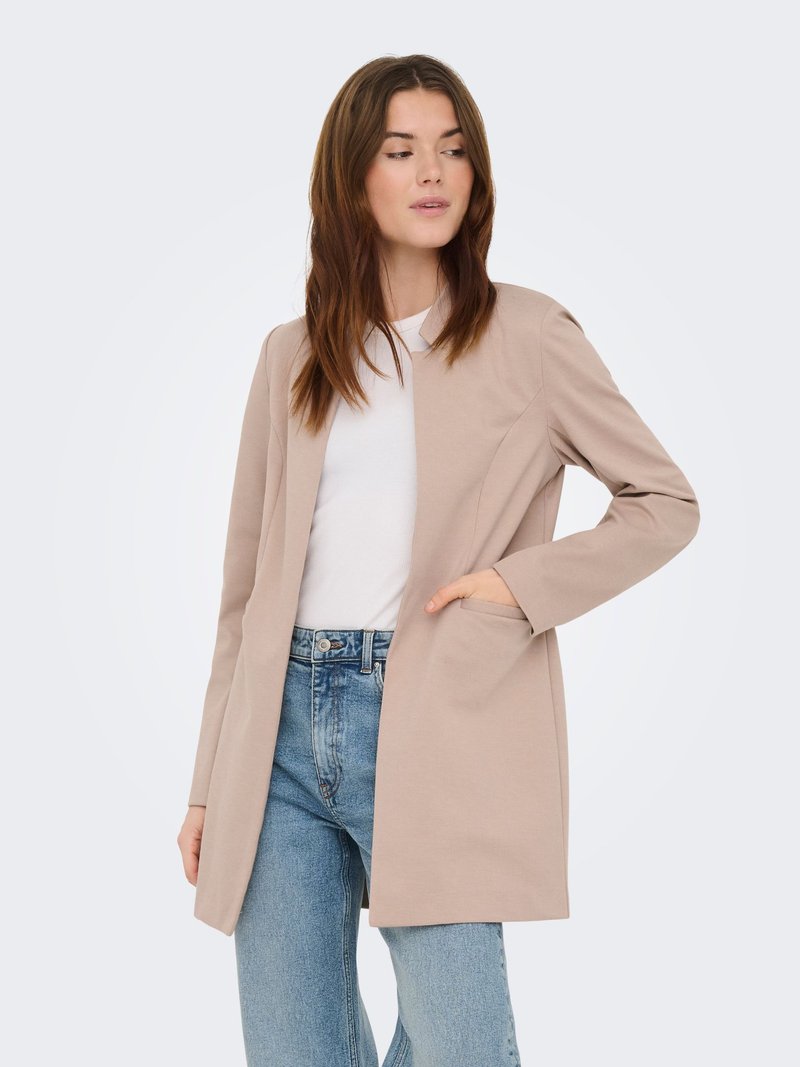 Blazer beige à ouverture avant, manches longues et texture lisse, avec des poches et une coupe cintrée, porté avec un jean clair.
